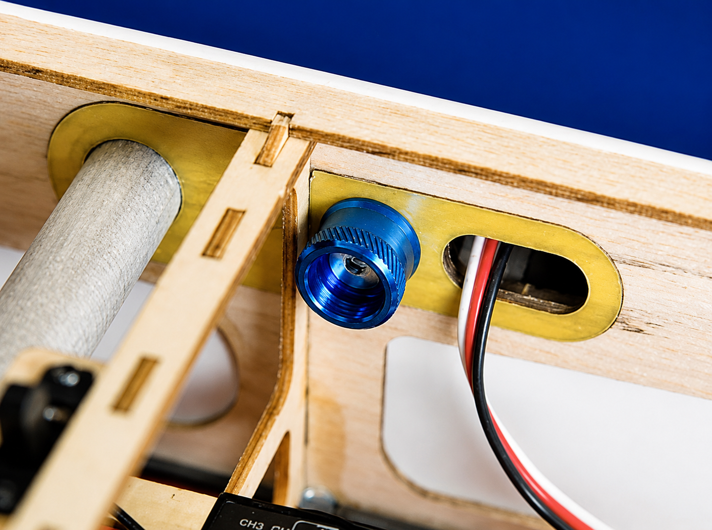 Close-up of a wooden mechanical device with a blue anodized metal connector, brass-colored brackets, and a white cylindrical rod, with red and black wires visible.